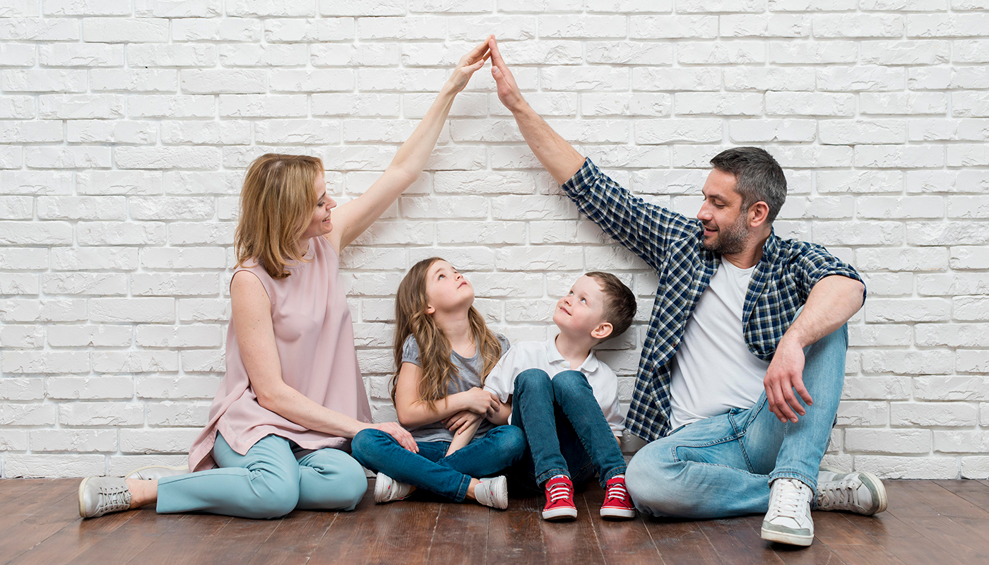 Familie sitzt gemeinsam vor einer Wand und die Arme der Eltern bilden ein Dach über den Kinderköpfen
