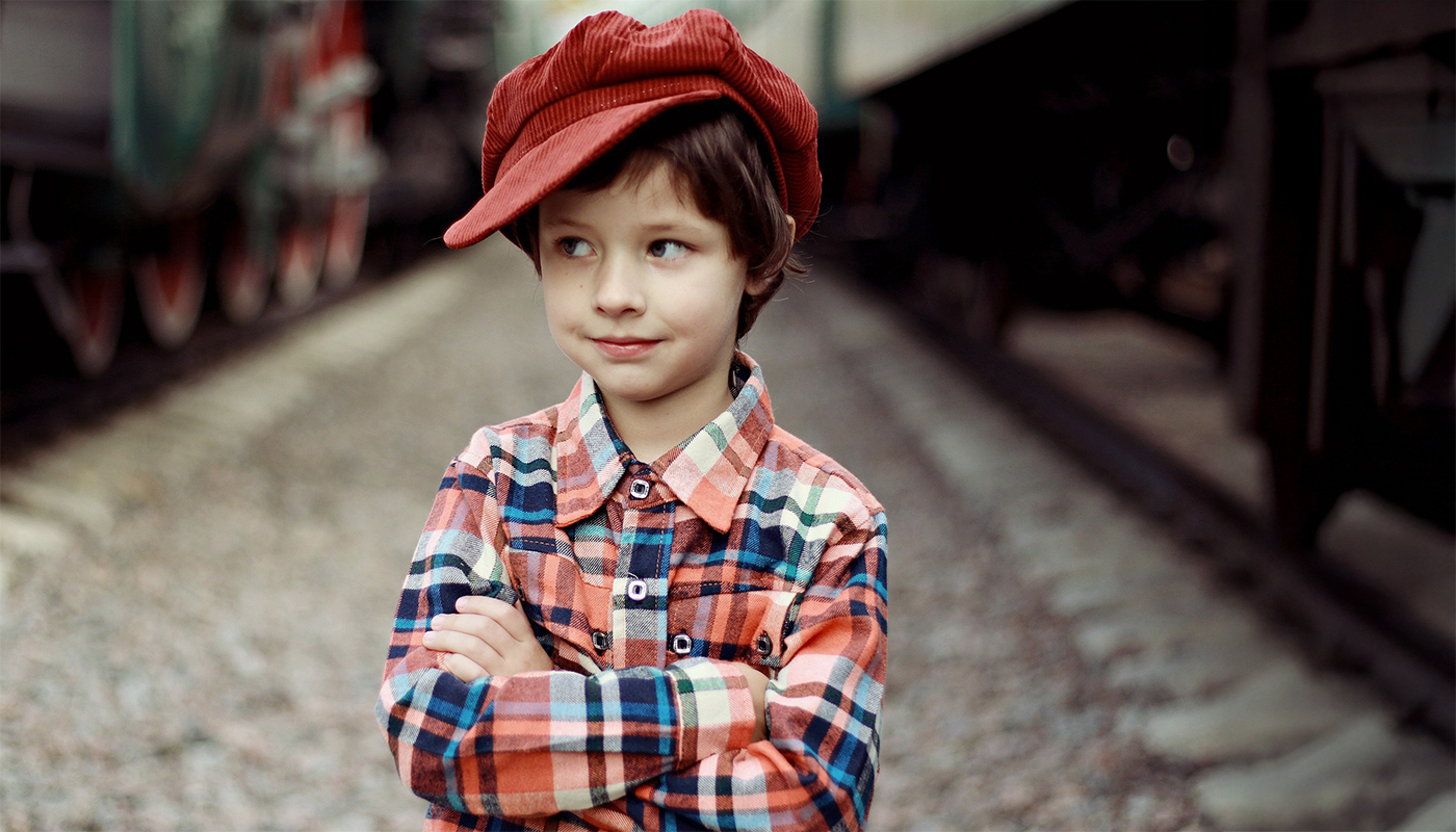 Kind mit roter Kordmütze und kariertem Hemd steht mit verschränkten Armen im Mittelpunkt; sein Blick zur Seite gekehrt;